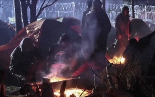 Ihmisiä kahden nuotion äärellä, taustalla telttoja ja piikkilanka-aita.