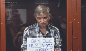 Aleksei Gorinov kuvattuna lasin takaa. Hän pitelee lappua, jossa on venäjänkielistä kirjoitusta.